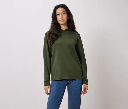 Eine Frau mit langen braunen Haaren posiert in einem olivgrünen Hoodie und blauen Jeans vor einem weißen Hintergrund.