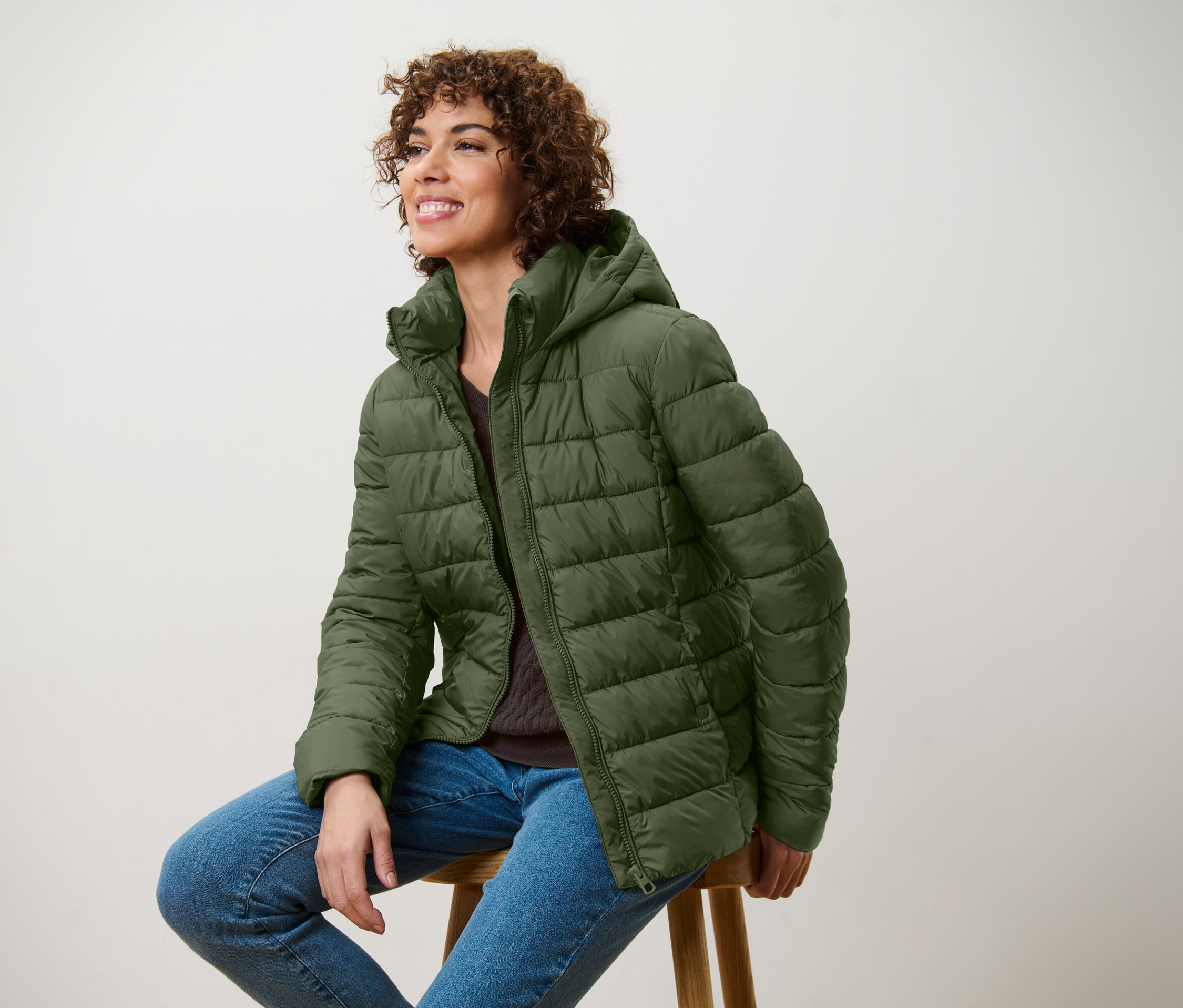 Lächelnde Frau sitzt auf einem Hocker und trägt eine olivgrüne Steppjacke und Jeans.