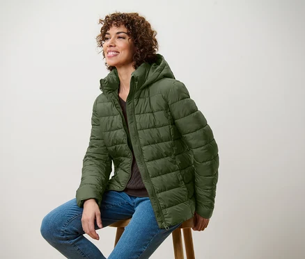 Lächelnde Frau sitzt auf einem Hocker und trägt eine olivgrüne Steppjacke und Jeans.