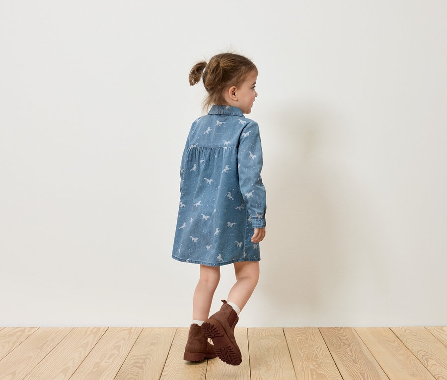 Mädchen in Jeanskleid mit Pferdeaufdruck und braunen Stiefeln steht seitlich zu einer weißen Wand.