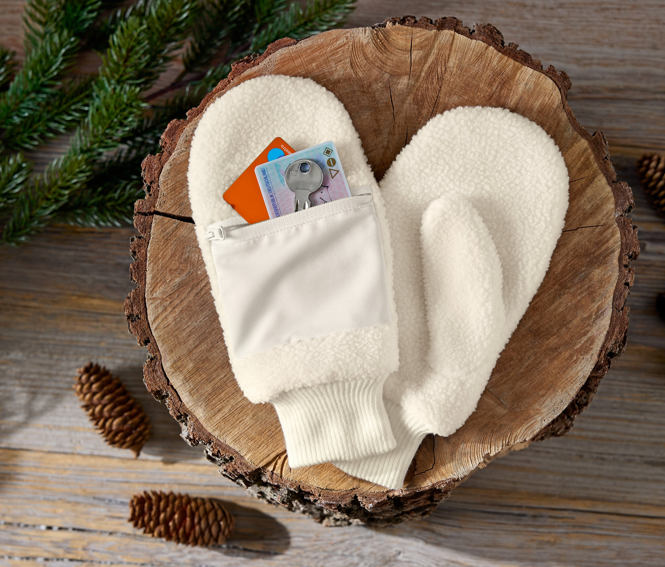 Cremefarbene Fäustlinge mit Reißverschlusstasche auf einem Holzuntergrund mit Tannenzapfen und Zweigen.