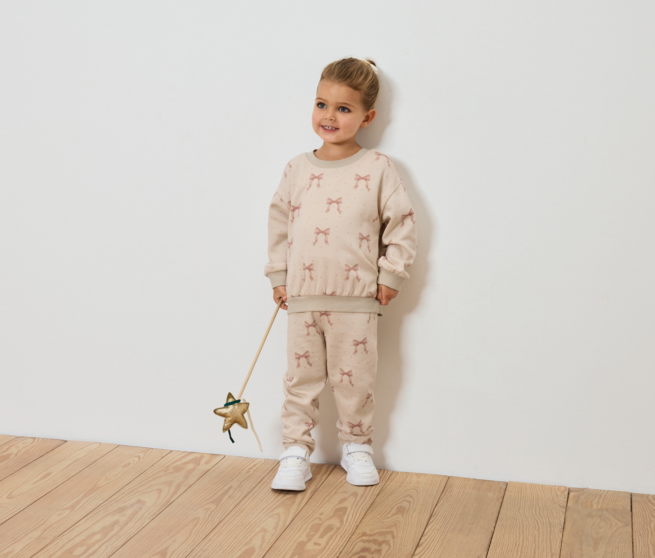 Ein lächelndes kleines Mädchen hält einen sternförmigen Zauberstab und trägt ein beiges Outfit mit Schleifenmuster.