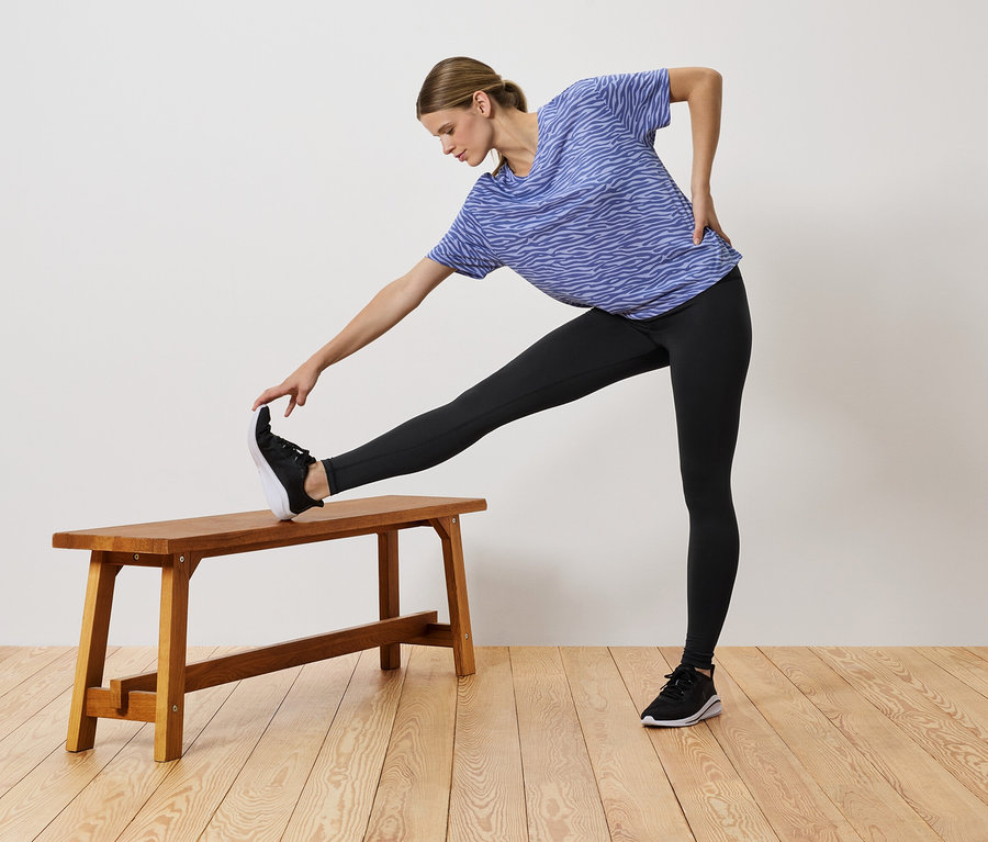 Frau in blauem T-Shirt und schwarzen Leggings dehnt sich mit einem Bein auf einer Holzbank.