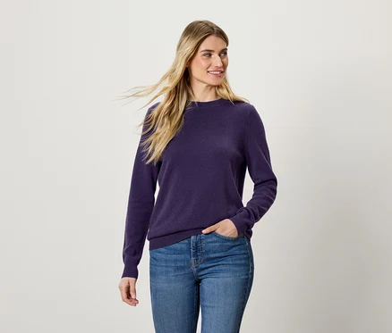 Frau mit blonden Haaren trägt einen violetten Pullover und blaue Jeans.