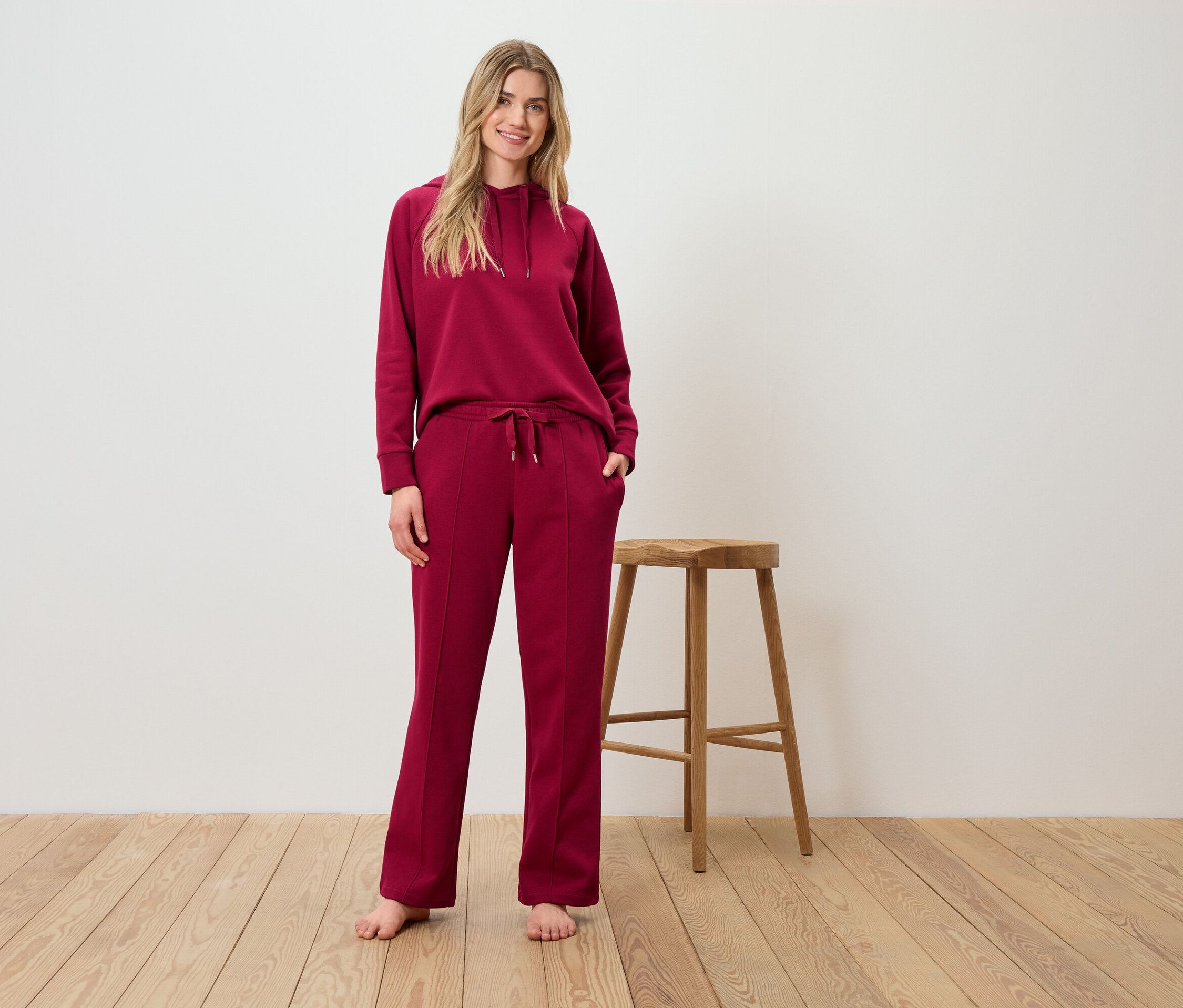 Eine Frau posiert in einem burgunderfarbenen Hoodie- und Hosenanzug vor einem Holzhocker.