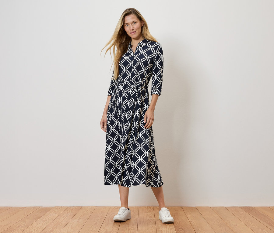 Frau steht und trägt ein marineblaues gemustertes Kleid und weiße Turnschuhe.