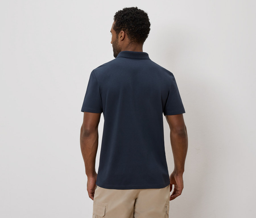 Rückansicht eines Mannes mit dunklem Haar, der ein marineblaues Poloshirt und beigefarbene Cargoshorts vor einem schlichten Hintergrund trägt.