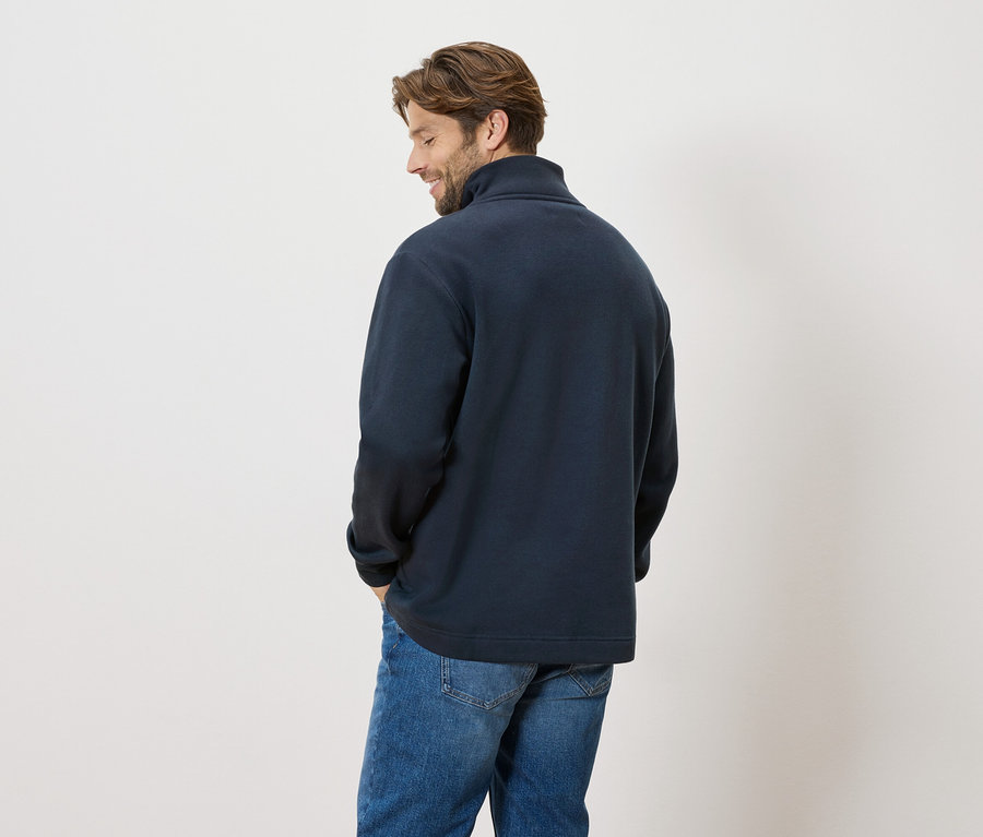 Ein Mann steht mit dem Rücken zur Kamera, er trägt einen dunkelblauen Pullover und Jeans.