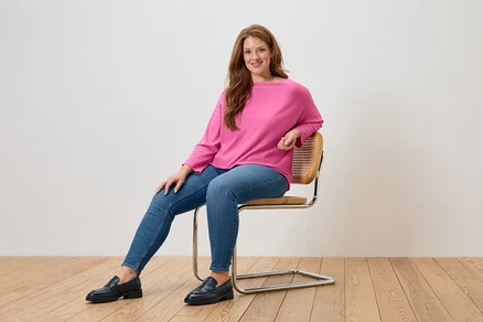 Eine Frau sitzt auf einem Stuhl und trägt einen pinkfarbenen Pullover, blaue Jeans und schwarze Loafer.
