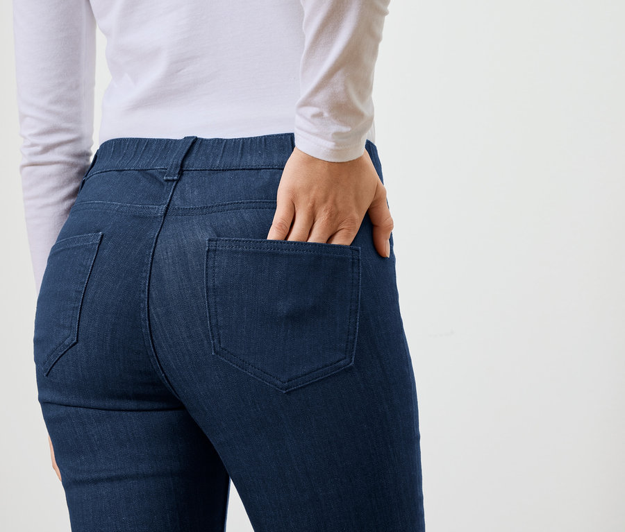 Frau mit Hand in der Gesäßtasche einer blauen Jeans, dazu ein weißes, langärmeliges Oberteil.
