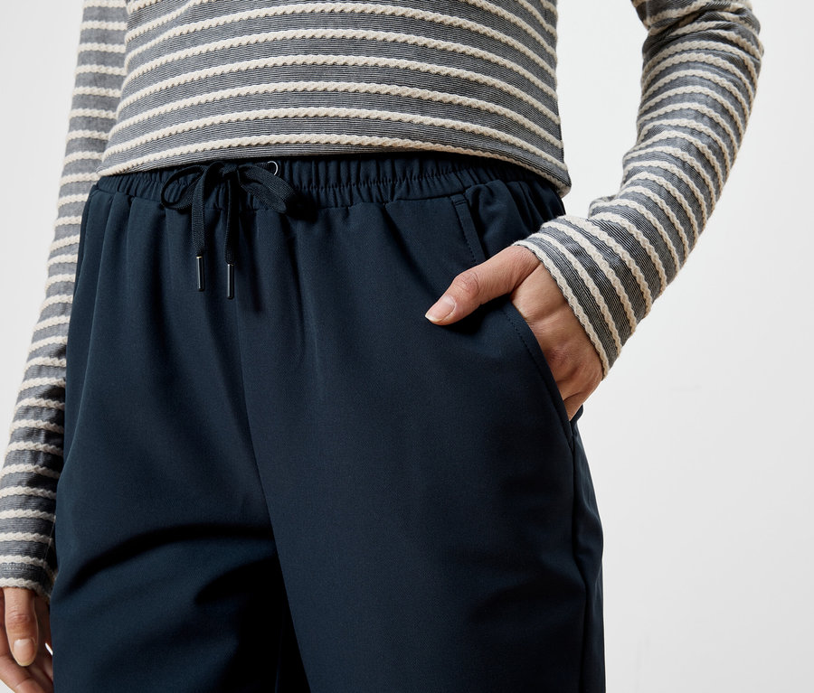 Frau mit gestreiftem Oberteil und marineblauer Hose mit Kordelzug, Hand in der Hosentasche.