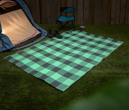 Blaues Zelt steht neben einer grün-grau karierten Picknickdecke auf Gras. Zwei Stiefel und ein blauer Klappstuhl stehen auf der Decke.