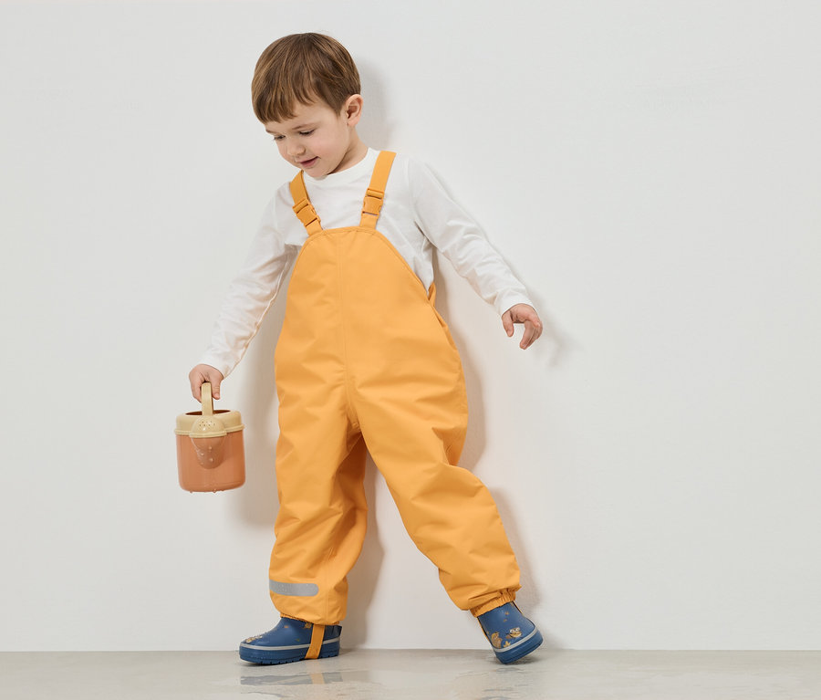 Ein Junge in gelber Latzhose und blauen Gummistiefeln hält eine Gießkanne vor einer weißen Wand.