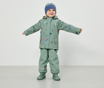 Ein Kleinkind in hellgrüner Regenbekleidung mit Dinosaurier-Muster und blauer Mütze steht mit ausgestreckten Armen vor einem weißen Hintergrund.
