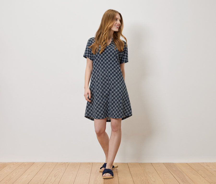 Eine Frau steht vor einer weißen Wand und trägt ein blaues Kleid mit Allover-Muster.