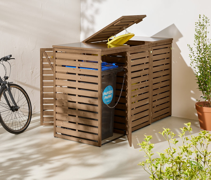 Drei braune Mülltonnenboxen stehen draußen, eine davon mit geöffneter Tür und Mülltonne. Daneben ein Fahrrad und eine Pflanze im Topf.