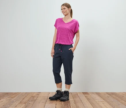 Eine Frau steht mit den Händen in den Taschen vor einer weißen Wand. Sie trägt ein pinkfarbenes T-Shirt, blaue Dreiviertelhosen und schwarze Wanderschuhe.