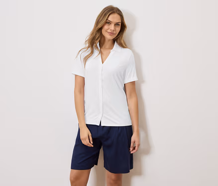 Eine Frau steht vor einer Wand und trägt ein weißes Hemd und blaue Shorts.
