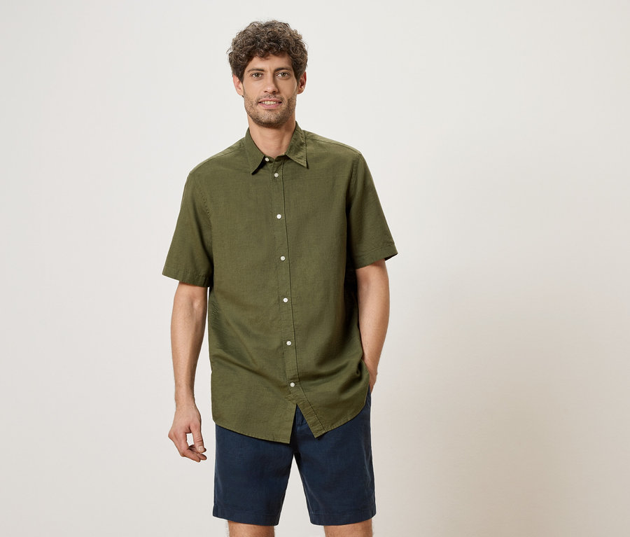 Ein Mann mit lockigem Haar steht und trägt ein olivgrünes Hemd mit kurzen Ärmeln und blaue Shorts.