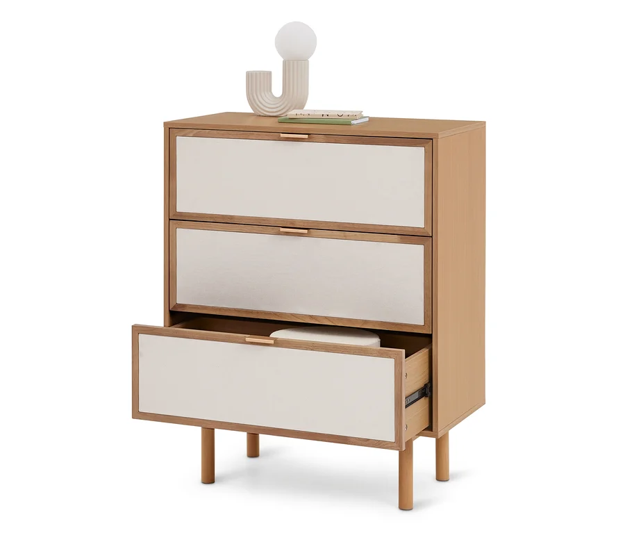 Helle Holzkommode mit drei cremefarbenen Stoffschubladen und goldenen Griffen. Die unterste Schublade ist geöffnet und zeigt ein weißes Objekt. Eine Dekoleuchte und Bücher stehen oben.
