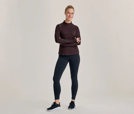 Eine lächelnde Frau steht mit verschränkten Armen, trägt ein dunkles langärmeliges Halb-Zip-Oberteil, dunkle Leggings und schwarz-weiße Sportschuhe.