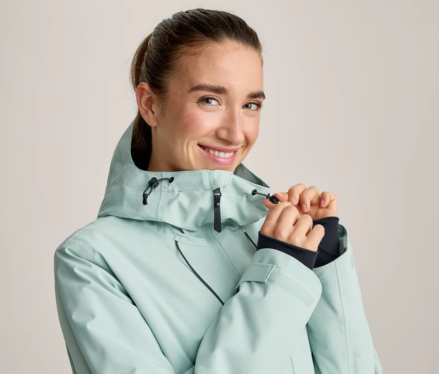 Eine lächelnde Frau mit zurückgebundenen Haaren, die die Kapuzenkordel ihrer hellgrünen Jacke mit beiden Händen anpasst.