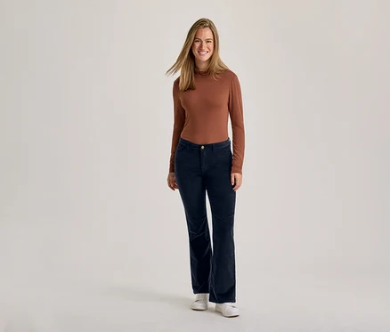 Eine lächelnde Frau steht in einem braunen Langarmshirt, dunkelblauen Cordhosen und weißen Turnschuhen.