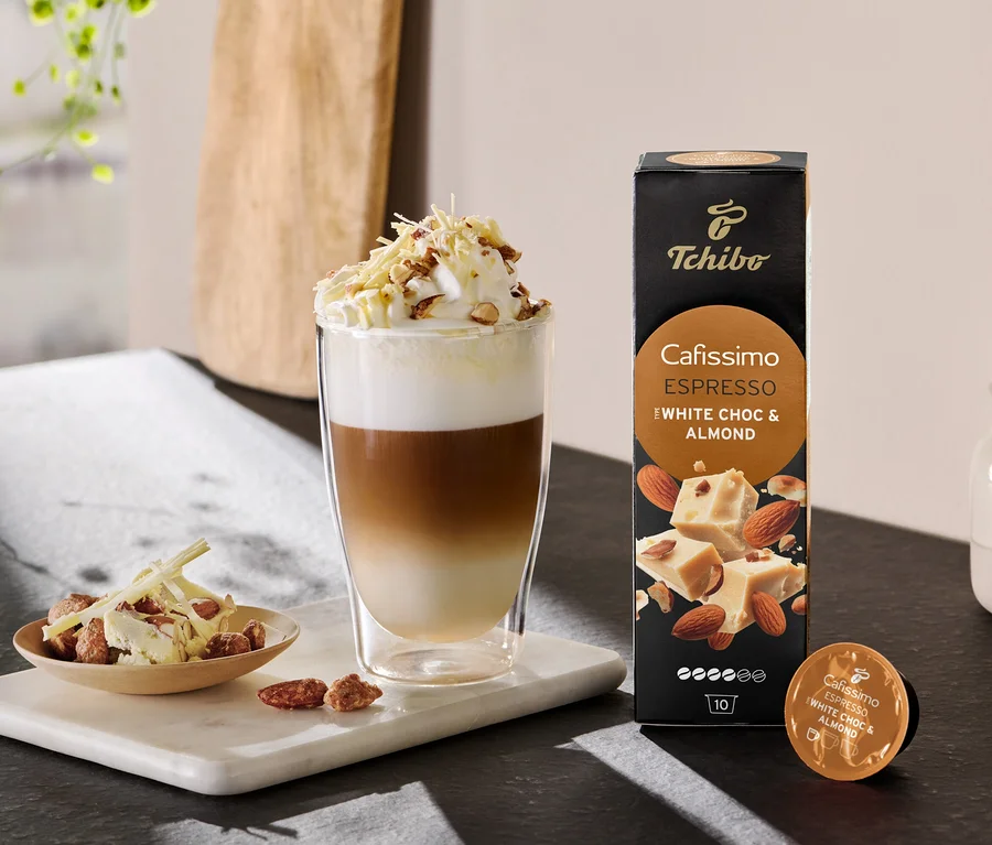 Geschichteter Latte mit Schlagsahne und Streuseln neben einer Packung Cafissimo Flavoured Espresso - White Choc & Almond.