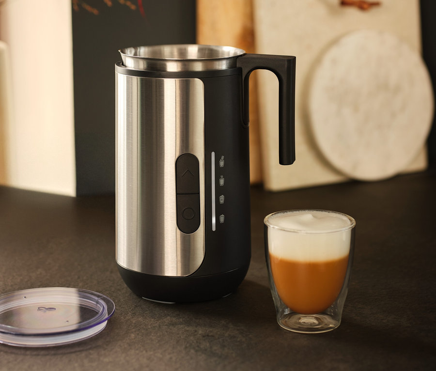 Ein Milchaufschäumer aus Edelstahl steht neben einem Glas Kaffee mit Milchschaum.