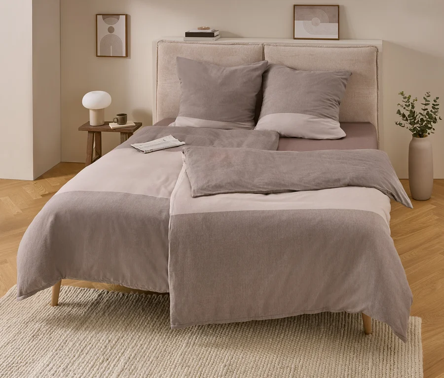Schlafzimmer mit Bett und hellbraun-taupe gestreifter Flanell-Bettwäsche. Links ein Holztisch mit Lampe und Tasse, rechts eine Vase mit Grünpflanzen. Eine Zeitung liegt auf dem Bett.