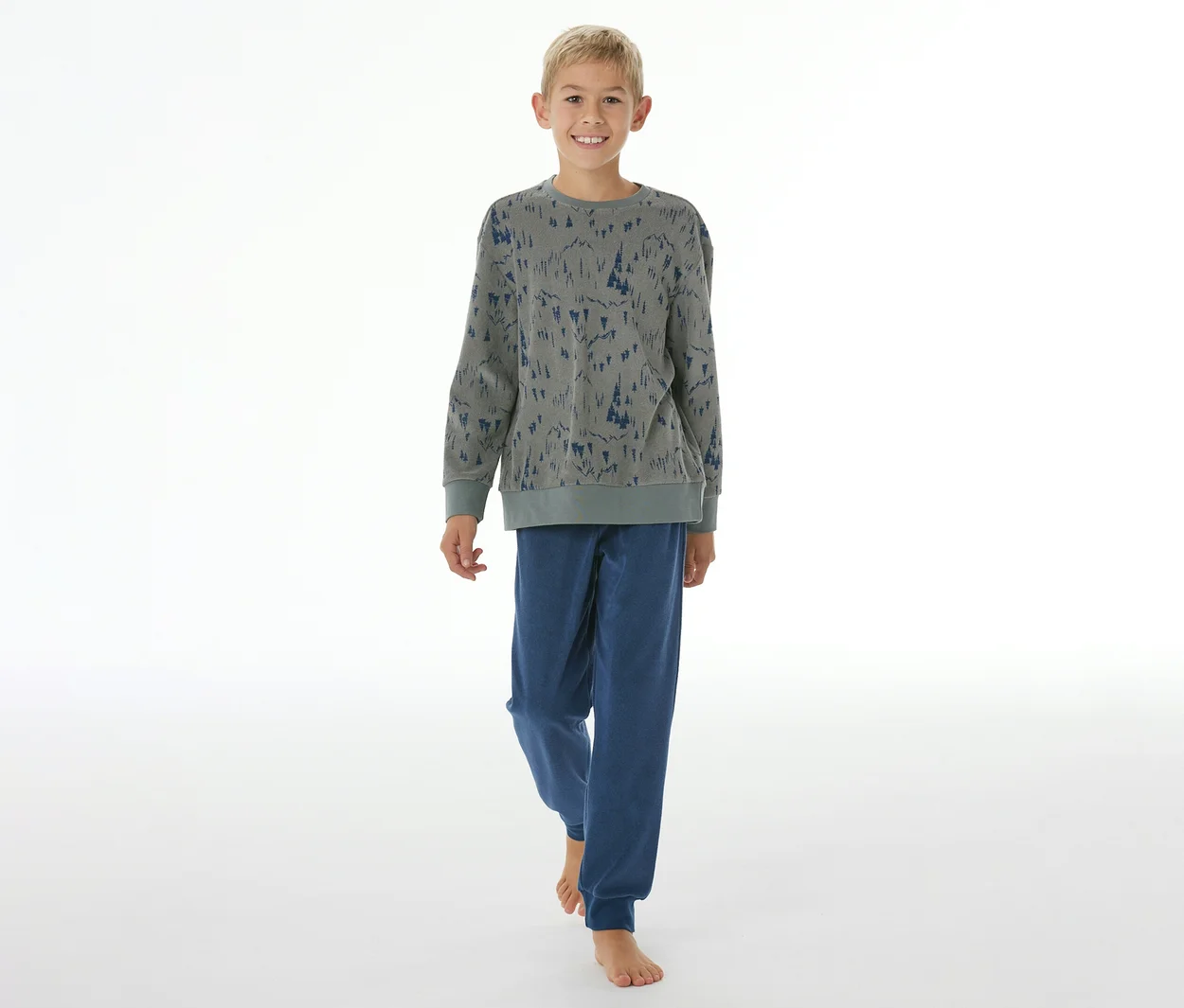 Lächelnder Junge in SCHIESSER Kinder-Pyjama mit Bündchen: graues Oberteil mit blauem Baummuster, blaue Hose. Er steht barfuß.