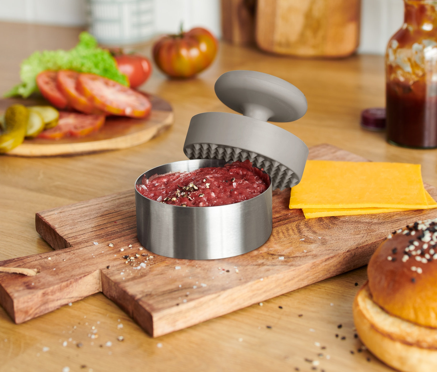 Auf einem Holzbrett liegen ein Burger-Brötchen, Käse und eine geöffnete Burgerpresse mit Fleisch.