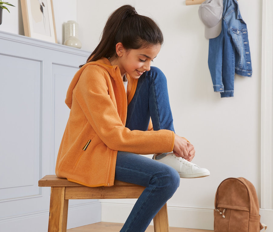 Ein Mädchen sitzt auf einem Holzhocker und bindet sich einen weißen Schuh zu. Sie trägt eine gelb melierte Strickfleece-Jacke.