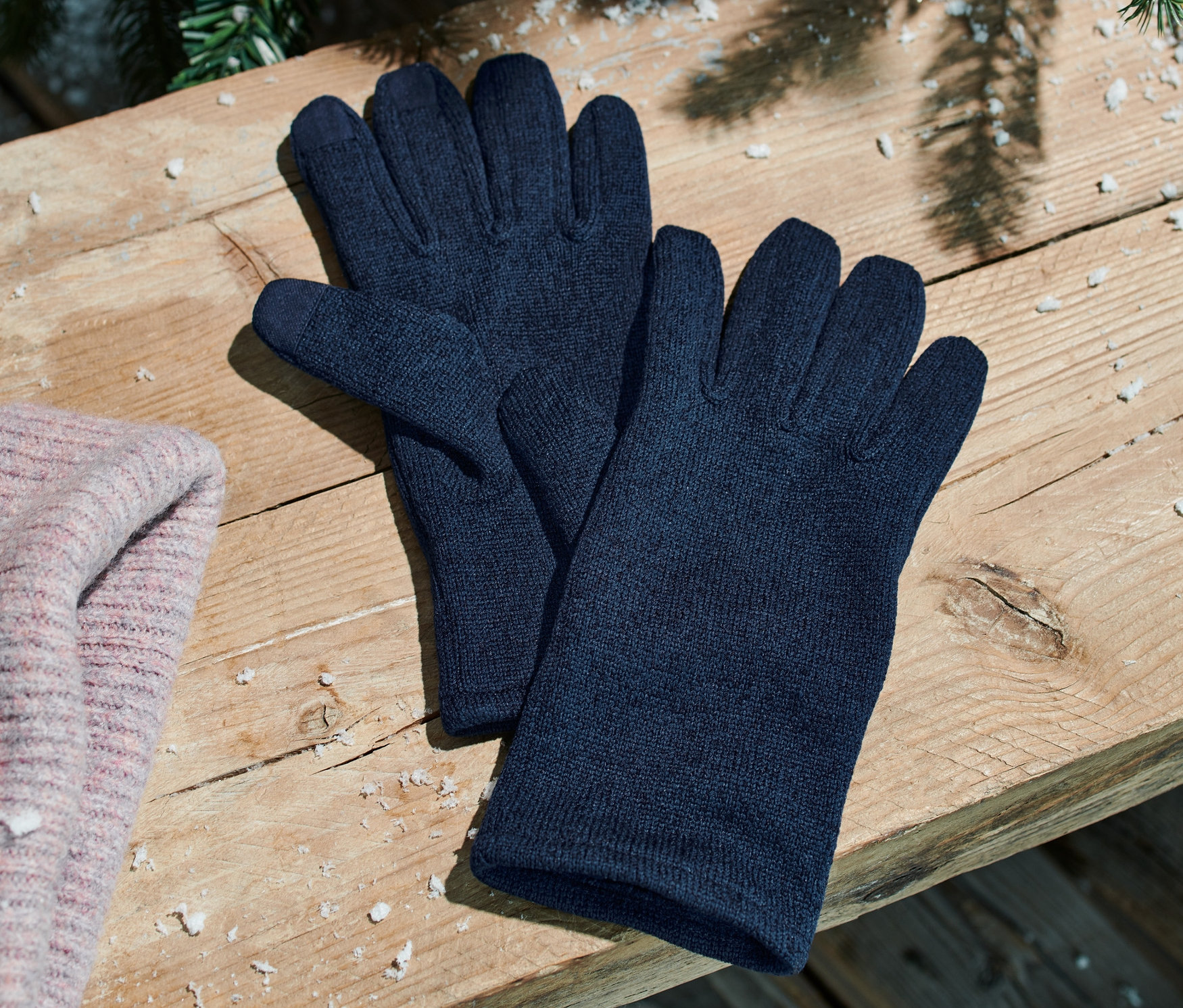 Blaue Strickfleece-Handschuhe liegen neben einer rosa Mütze auf einer Holzbank.