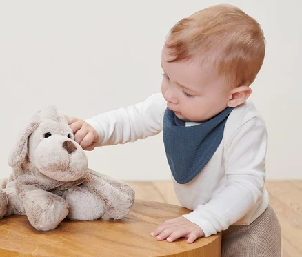 Ein Baby spielt mit einem Plüschhund auf einem Holztisch und trägt ein 2 Baby-Kleinkind-Musselin-Halstuch.