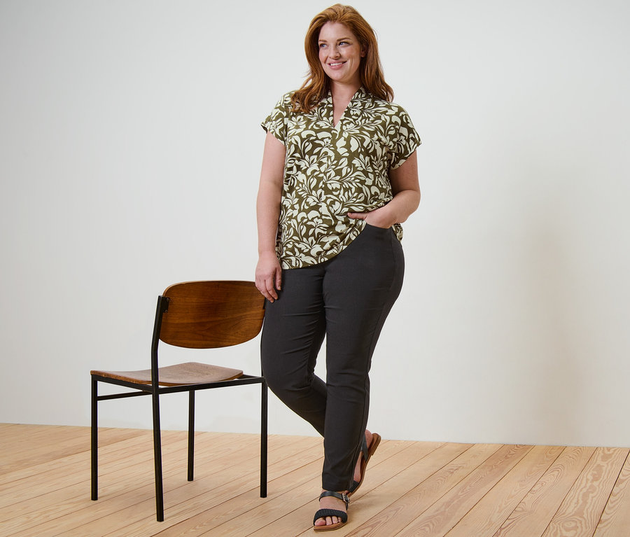 Frau posiert stehend in einem bedruckten Blusenshirt mit V-Ausschnitt und einer schwarzen Bengalin-Hose, die Hand in der Hosentasche, neben ihr steht ein Stuhl.