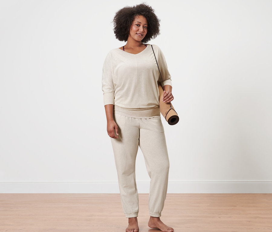 Eine Frau steht und hält eine Yogamatte. Sie trägt ein beiges Yogashirt und eine Sweathose.
