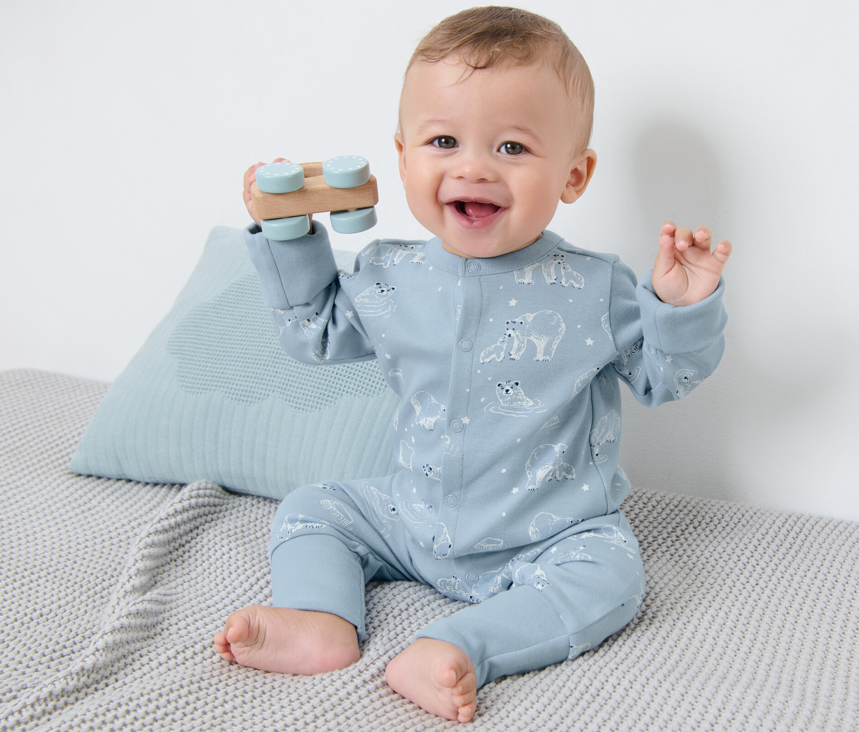 Lächelnder Säugling sitzt auf einer grauen Strickdecke und spielt mit einem Holzspielzeug. Er trägt einen blauen Baby-Pyjama mit Eisbärenmuster.