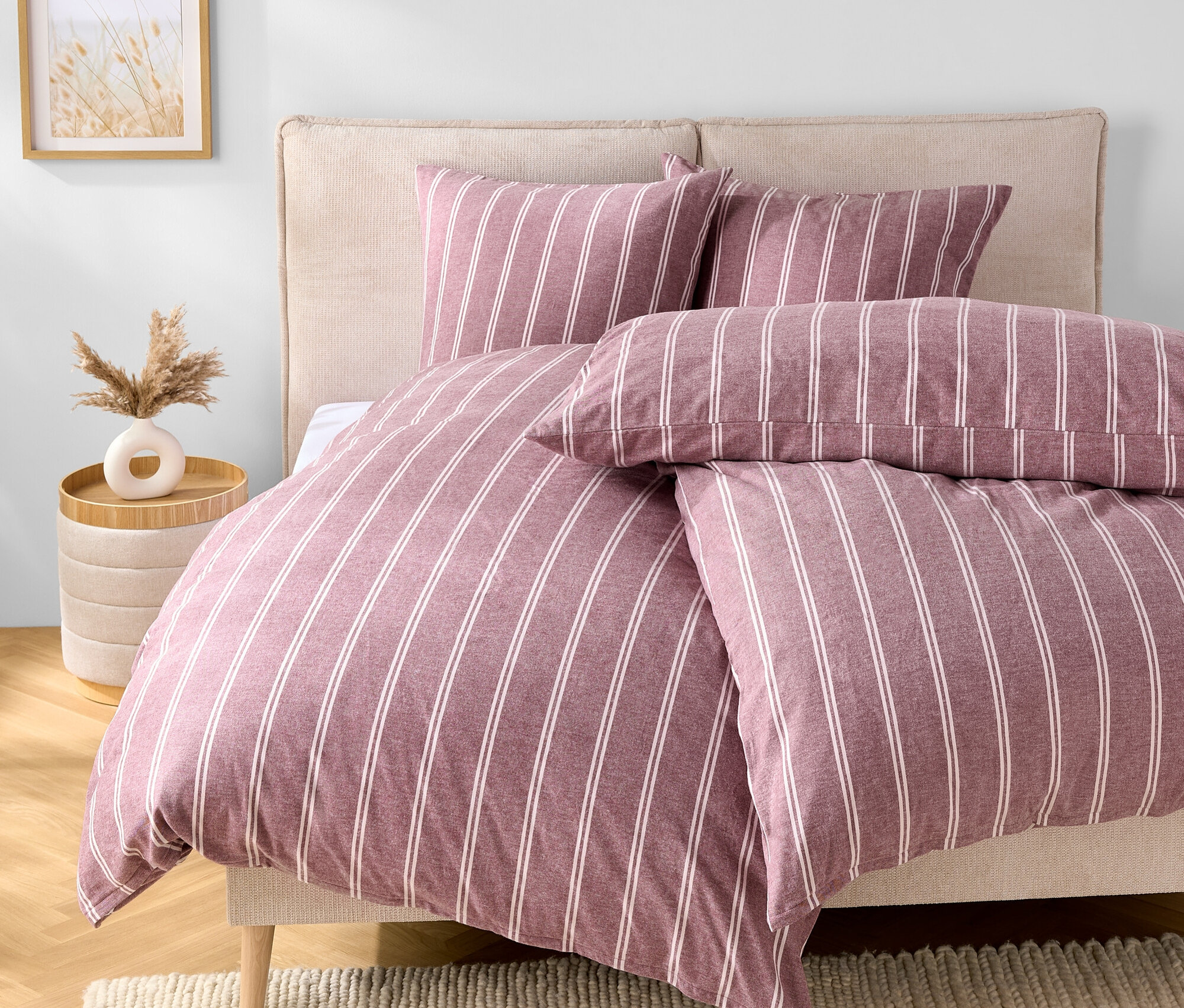 Schlafzimmer mit einem Bett mit lila-weiß gestreifter Bettwäsche, zwei Kissen und einem beigen Kopfteil. Neben dem Bett steht ein Tisch mit einer Vase.