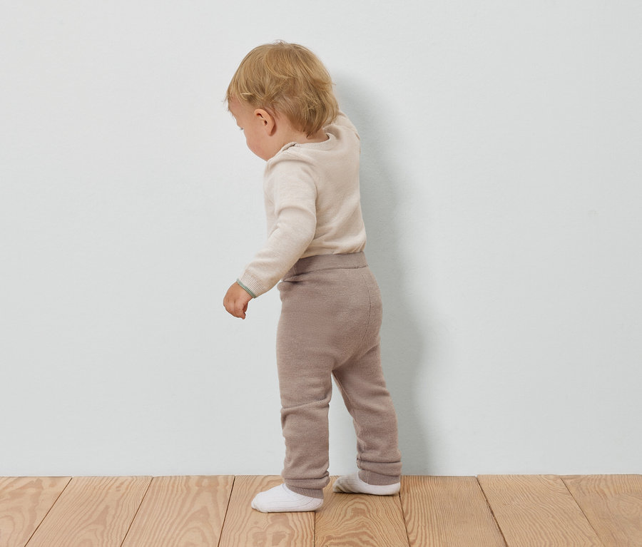 Kleinkind in cremefarbenem Pullover und brauner Hose steht mit dem Rücken zu einer weißen Wand.