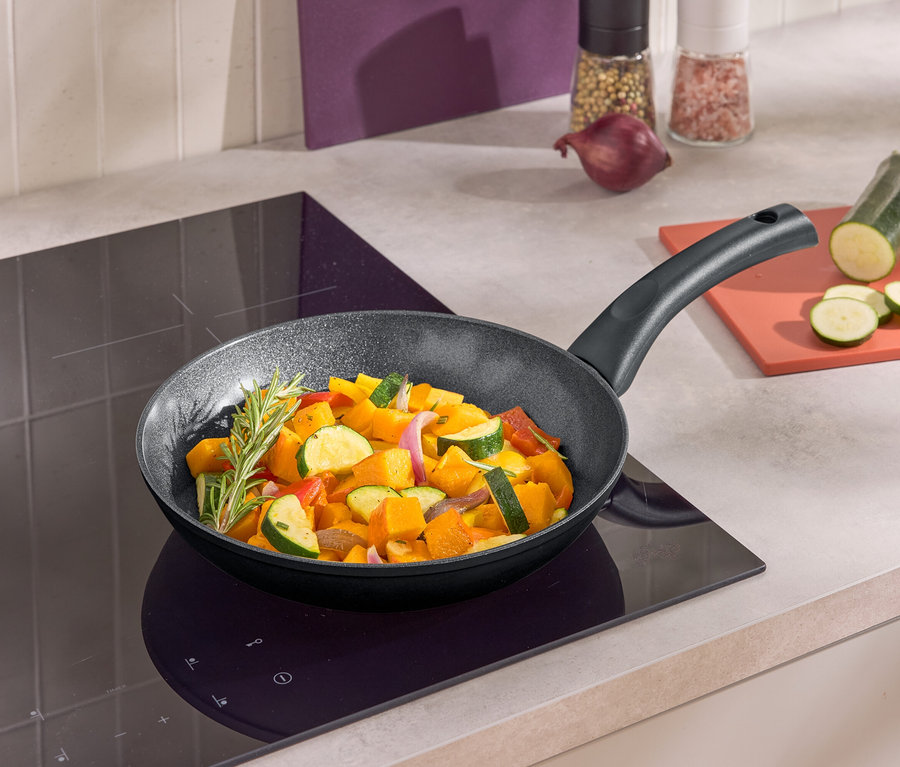 Eine Bratpfanne mit gebratenem Gemüse auf einem Induktionskochfeld. Neben der Pfanne liegt eine geschnittene Zucchini auf einem Brettchen.