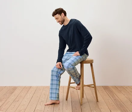 Mann sitzt auf einem Holzhocker, trägt einen dunkelblauen Schlafanzug und eine blau karierte Hose.
