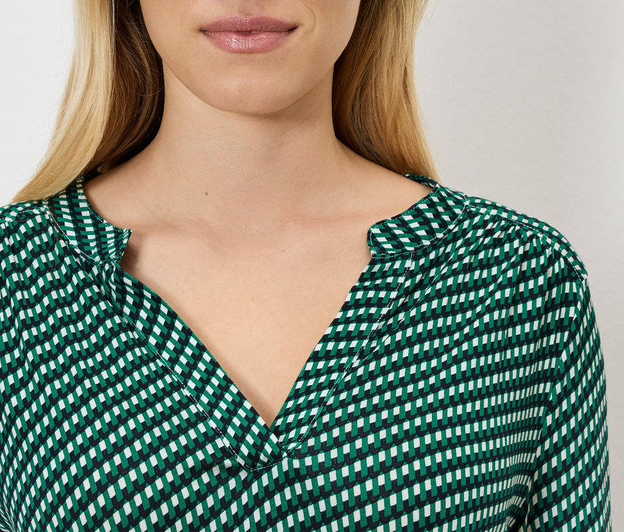 Nahaufnahme einer Frau mit blonden Haaren, die eine grün-weiß gemusterte Bluse trägt.