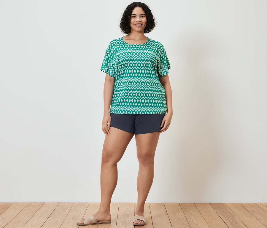 Frau posiert in einem grünen, bedruckten Oberteil und dunkelblauen Shorts. Sie trägt auch Sandalen.