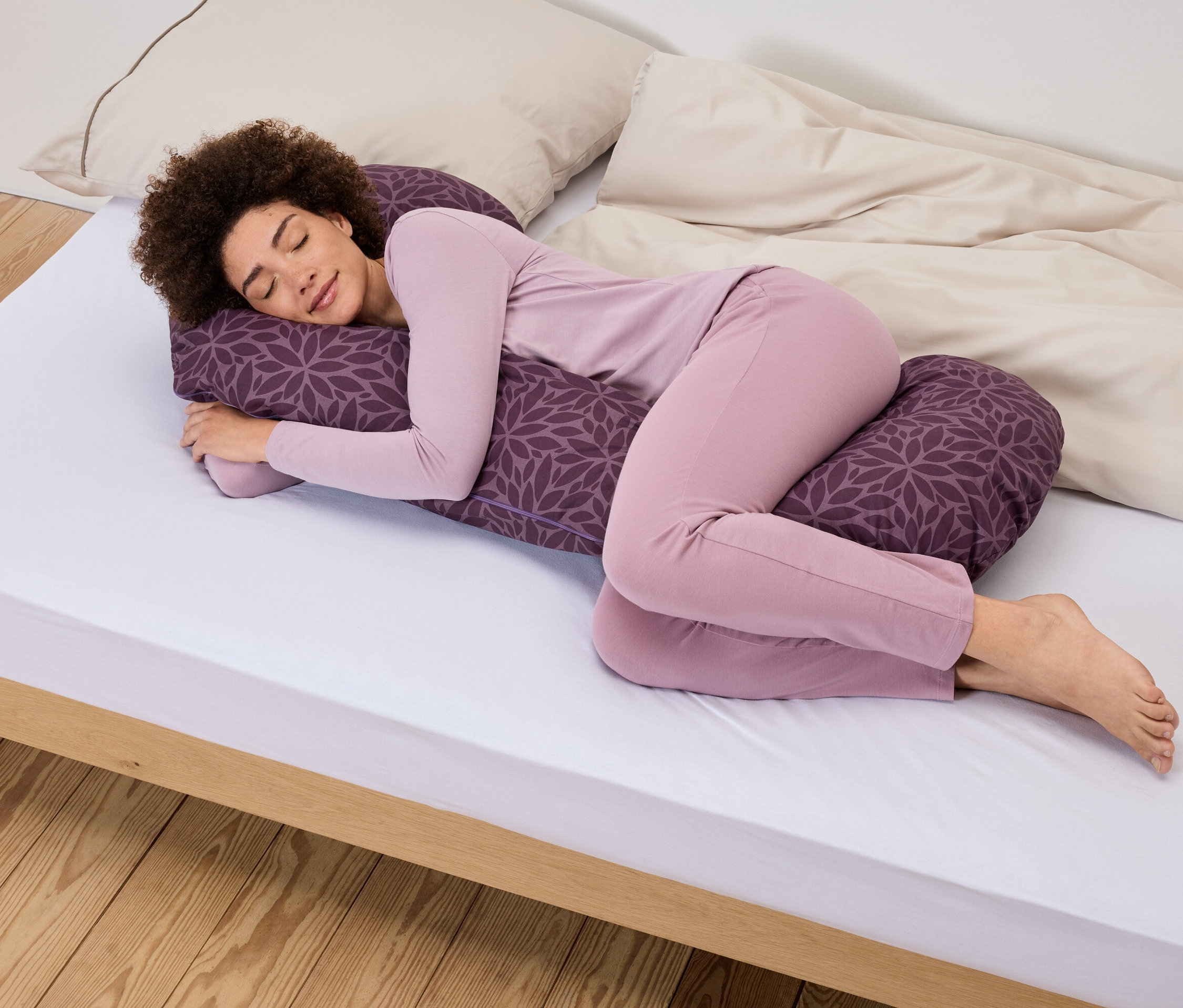 Eine Frau mit Afro-Frisur liegt seitlich auf einem weißen Bett und umarmt ein lila Kissen mit Blumenmuster.