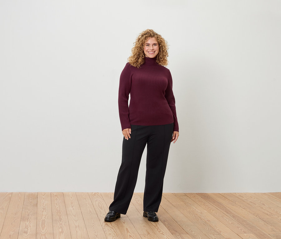 Eine Frau mit lockigem Haar steht und lächelt in einem burgunderroten Rollkragenpullover und einer schwarzen Hose.