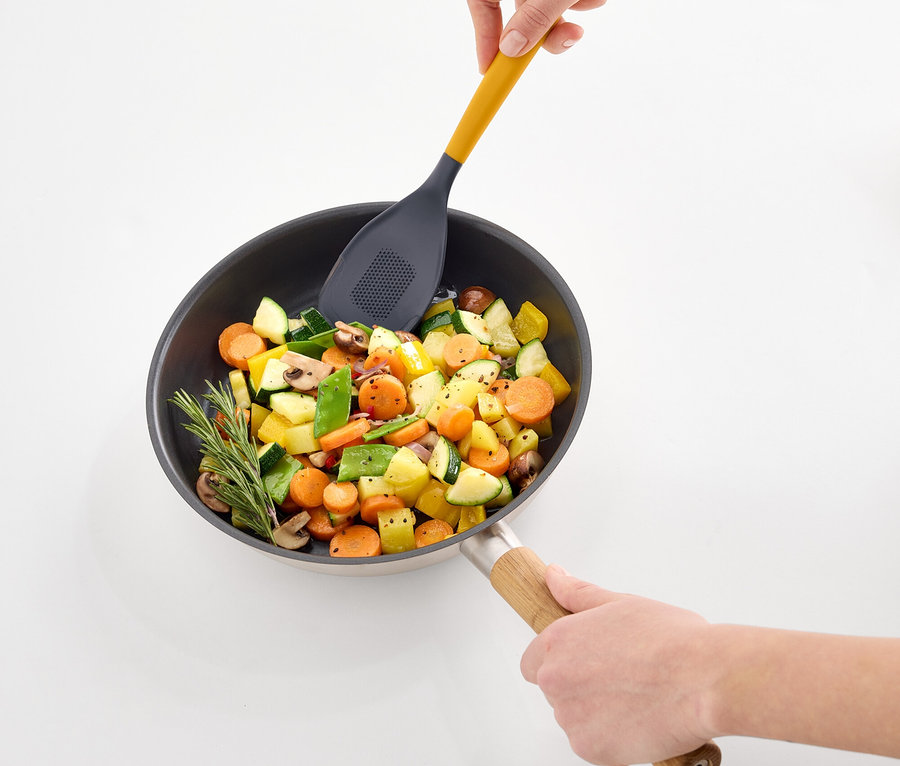 Hände verrühren Gemüse in einer Bratpfanne mit einem gelben, gelochten Pfannenwender.