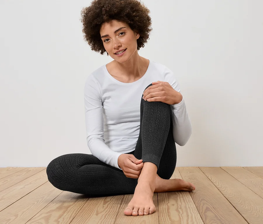 Eine Frau sitzt auf einem Holzboden und trägt graue Leggings und ein weißes Langarmshirt.