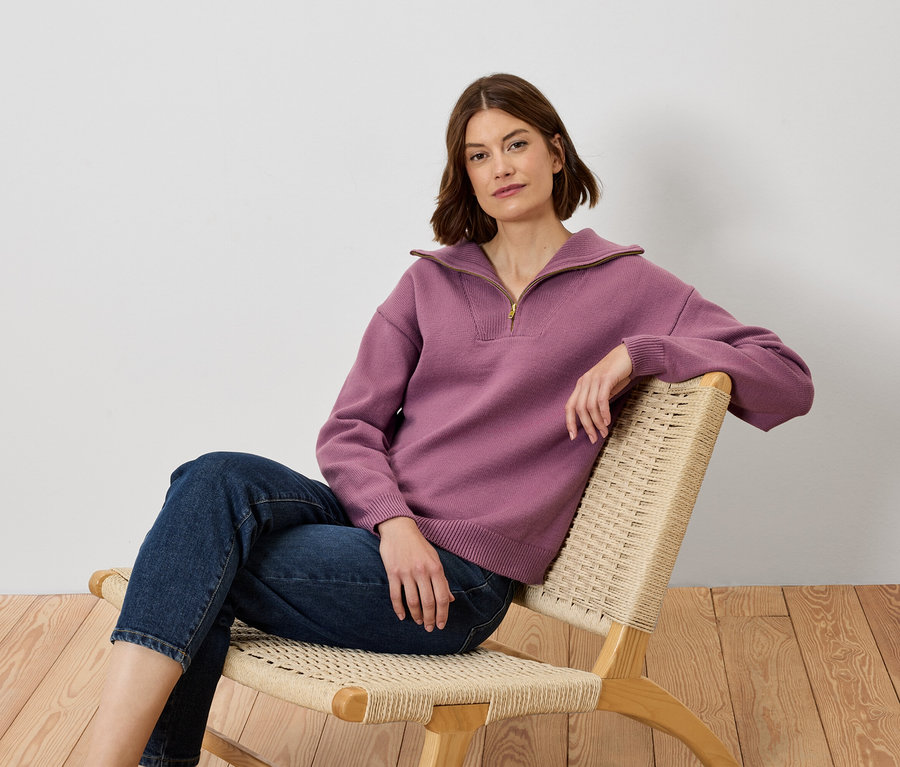 Frau sitzt in einem Stuhl, trägt einen lila Pullover und Jeans.