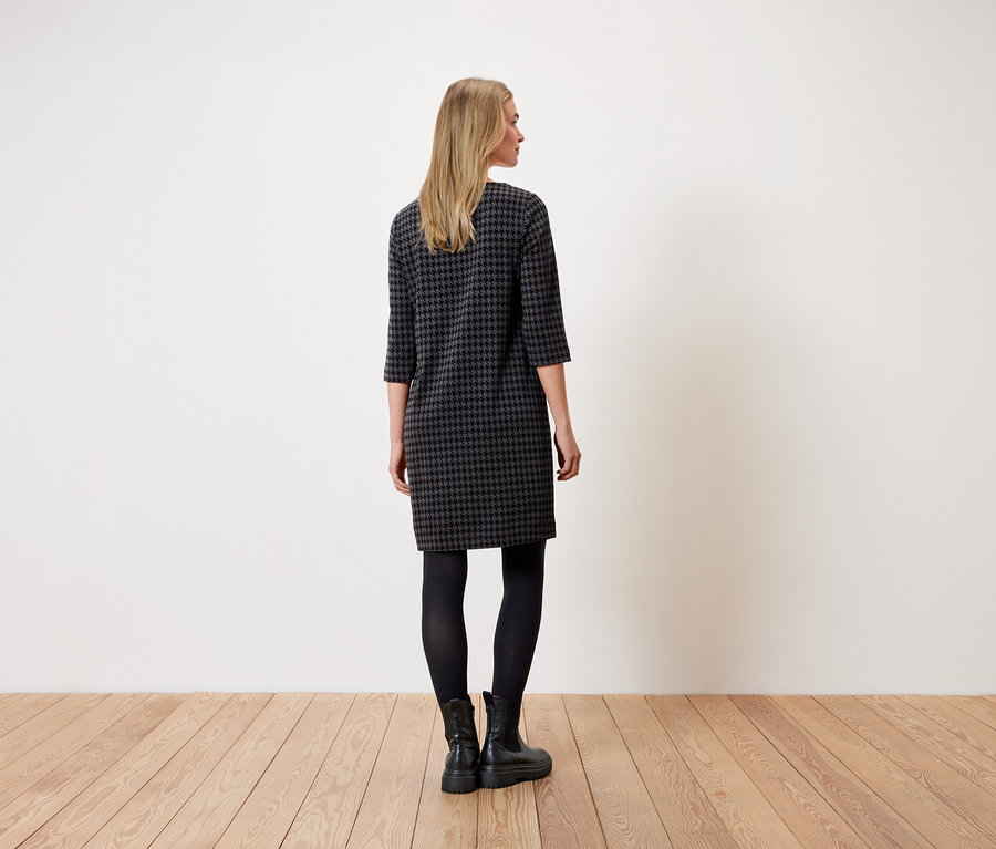 Eine Frau steht mit dem Rücken zur Kamera und trägt ein Hahnentrittmuster-Kleid, schwarze Leggings und schwarze Stiefel.